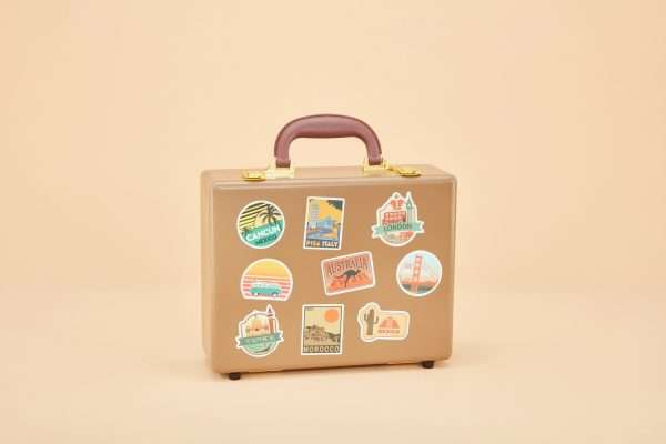 An urn that looks like a tan leather suitcase with gold latches and a brown handle. It is covered in stickers of cities around the world